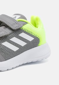 Zapatilla deportiva gris con parte superior de malla, acentos en verde neón, logo blanco y tira de Velcro ajustable. Suela blanca gruesa con surcos texturizados.
