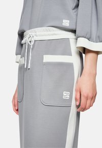 Graues Sportoutfit mit einem Oberteil mit voluminösen Ärmeln und einer Hose mit kontrastierenden weißen Akzenten und Taschen. Weiche, glatte Stofftextur.