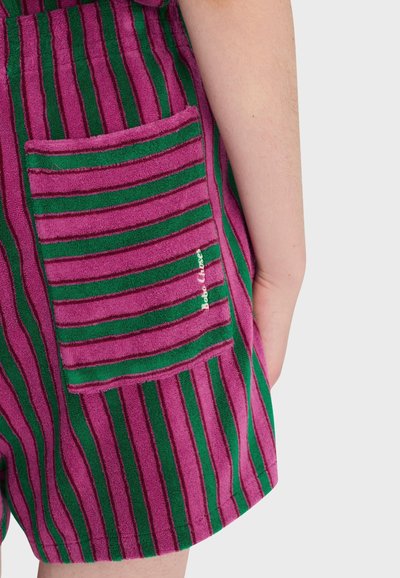 Vue latérale d'une personne portant un short rayé rose et vert avec une poche carrée sur la hanche droite, montrant une partie de son bras sur un fond uni.