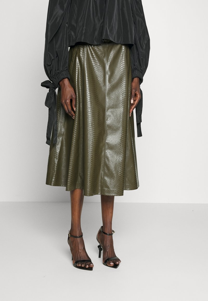 Personne portant une jupe midi olive brillante à motif peau de serpent, un chemisier noir à manches longues avec des poignets noués, et des talons noirs à bride cheville.