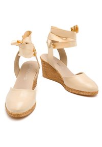 Beżowe sandały na koturnie z gładką skórzaną cholewką, okrągłym czubkiem i materiałowymi wiązaniami wokół kostki. Teksturowany, jutowy koturn wprowadza kontrast.