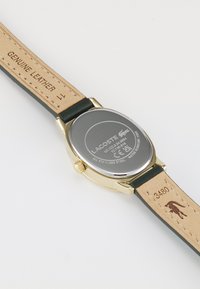 Montre dorée avec un boîtier rond en acier inoxydable. Elle comporte un bracelet en cuir véritable noir et des gravures au dos du boîtier et sur le bracelet.