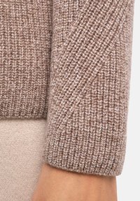 Beżowy sweter dziany z ukośną prążkowaną fakturą, miękki materiał i mankietowany rękaw. Wewnętrzna warstwa wydaje się jaśniejsza i gładka.