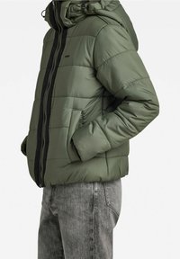 Zielona pikowana kurtka z kapturem, z czarnym zamkiem błyskawicznym i bocznymi kieszeniami. Materiał jest gładki, o lekkiej, pikowanej fakturze.