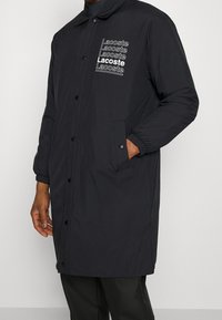 Černý dlouhý kabát z umělého materiálu, s knoflíkovým zapínáním, elastickými manžetami a více bílými logy "Lacoste" na přední straně.