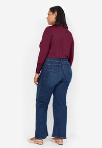 Haut à manches longues bourgogne avec des accents en dentelle, associé à un jean flare en denim bleu. Le mannequin porte des chaussures plates de couleur claire et a les cheveux noirs raides.
