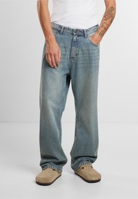 Világoskék bő szabású farmernadrág, elhalványult textúrával, öt zsebbel és gombos zárással, bézs slip-on lábbelivel párosítva.