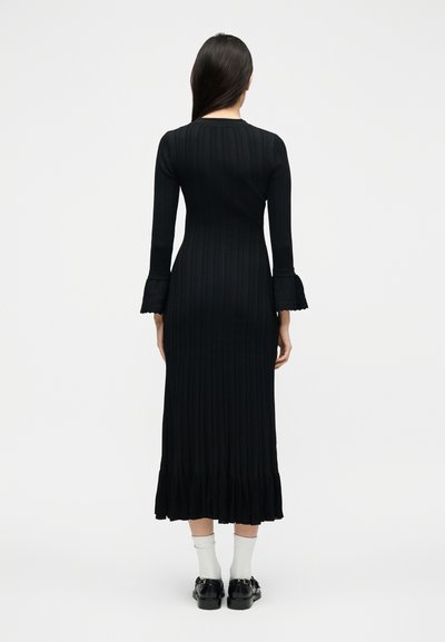 Γυναίκα με μακριά σκούρα μαλλιά που φοράει μαύρο ριμπ midi φόρεμα με φουσκωτά δαντελένια μανίκια και βολάν στο κάτω μέρος, λευκές κάλτσες και μαύρα μοκασίνια, γυρισμένη με την πλάτη.