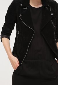 Schwarze Wildleder-Motorradjacke mit Reißverschluss vorne, Druckknopfdetails und umgeschlagenen Ärmeln. Über einem schwarzen Oberteil getragen, verfügt sie über Seitentaschen und eine taillierte Passform.