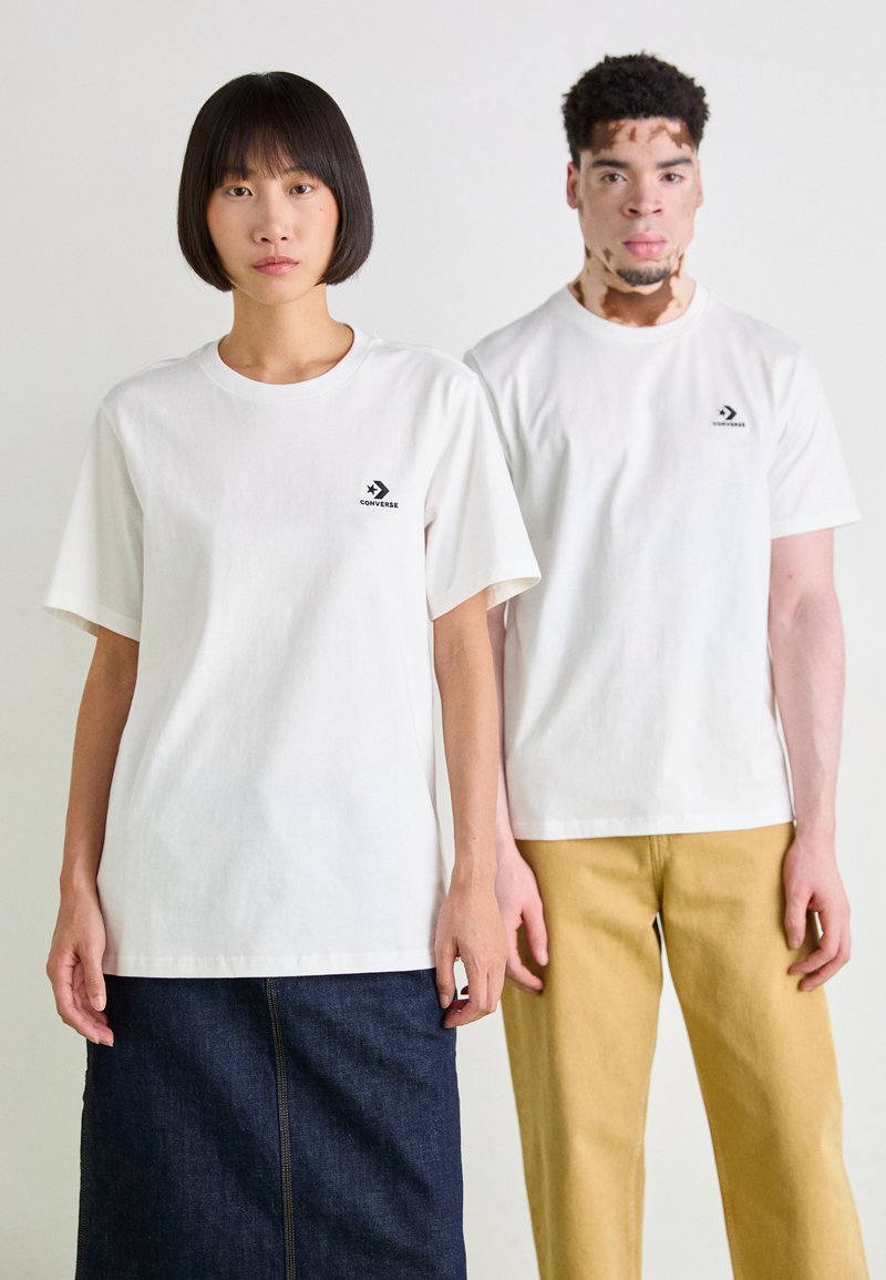 Dos personas llevan camisetas de algodón blancas con un logo negro de Converse. La mujer la combina con una falda de mezclilla oscura, mientras que el hombre usa pantalones beige claros.