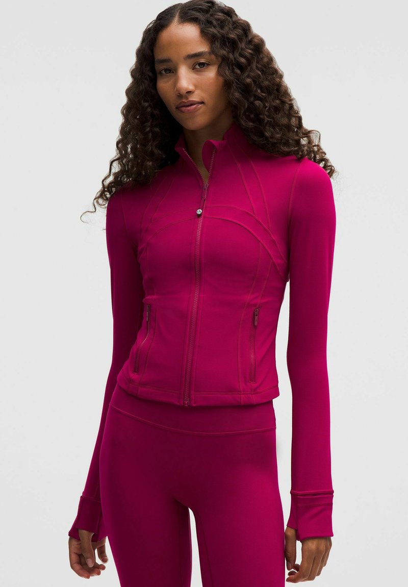 Kitsas magenta spordijakk kõrge kraega, esiüksene tõmblukk ja tasane õmblus. Omab pikki varrukaid koos sõrmeaukudega ja tõmmatavaid taskuid.