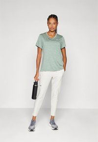 Πράσινο αθλητικό πουκάμισο από ελαφρύ ύφασμα, συνδυασμένο με λευκό στενό παντελόνι και γκρι παπούτσια. Μπουκάλι νερό στο χέρι. Ορατό το λογότυπο Under Armour.