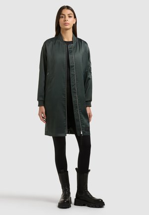 Frau steht vor einem schlichten hellen Hintergrund und trägt eine lange, dunkelgrüne Bomberjacke, schwarze Leggings und schwarze, klobige Stiefel.