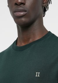 T-shirt vert en coton avec un col rond, présentant un petit détail brodé blanc "II" sur le côté gauche de la poitrine. Texture lisse.