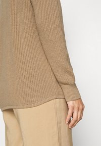 Beige sweater van wafelbreiwerk met geribbelde manchetten en zoom, met een losse pasvorm en zijsplitten, gecombineerd met lichtbeige broeken.