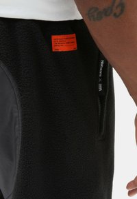 Sorte fleece joggers med en sidelomme, glat tekstur, grå stofdetaljer og et rødt mærke på bagsiden.