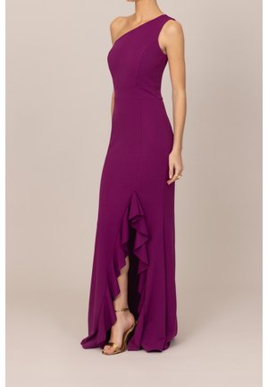 Femme portant une robe violette sans manches avec une bretelle sur une épaule et une fente avant à volants, associée à des sandales dorées à talons hauts.