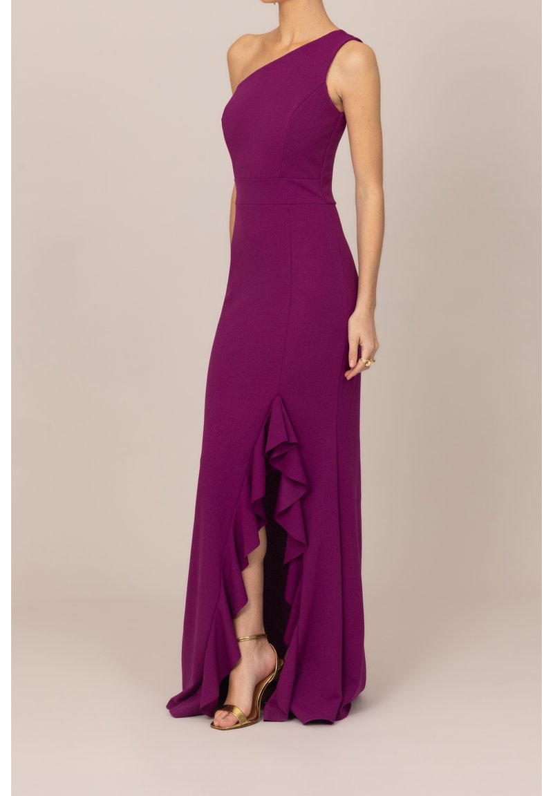 Femme portant une robe violette sans manches avec une bretelle sur une épaule et une fente avant à volants, associée à des sandales dorées à talons hauts.