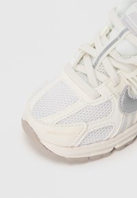 Artimas baltos tinklinės ir odinės sportinės avalynės kadras su smėlio spalvos guminiu padu ir šviesiai pilku Nike ženkliuku.
