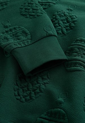 Dark green textured fabric with raised pinecone and ornament patterns and a close-up of a ribbed cuff edge.