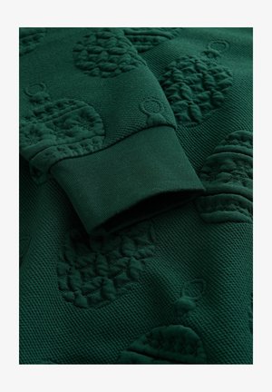 Tumši zaļš teksturēts audums ar izceltiem priedes konusveida un rotājumu rakstiem, kā arī tuvplāns ar ribotu piedurknes malu.