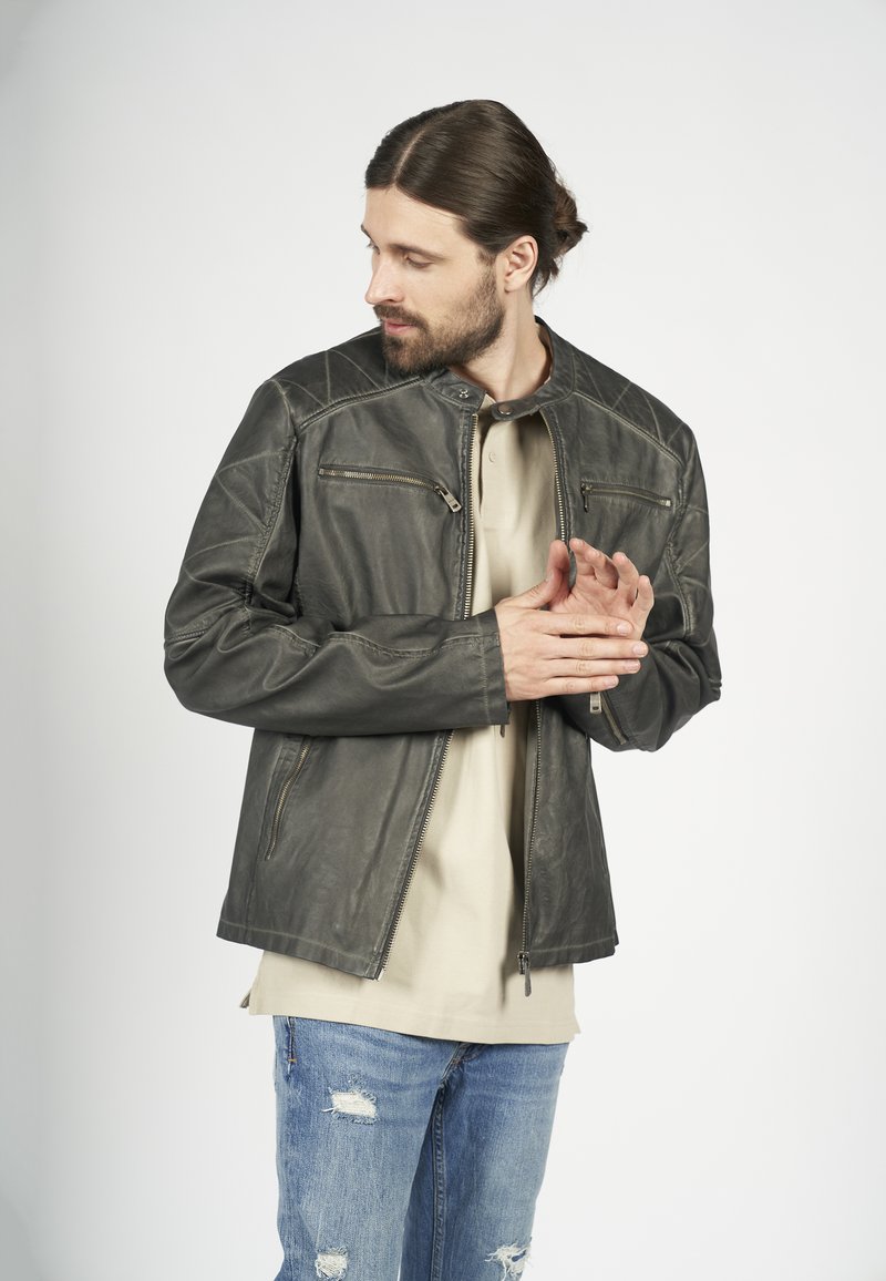 Mörkgrå läderjacka med hög krage, snedställd söm och två framfickor med dragkedja, som bärs över en beige t-shirt och blå jeans.