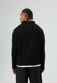 Zwart gebreid jasje met lange mouwen, brede kraag, gestructureerde stof en een losse pasvorm, gedragen over een wit overhemd en gecombineerd met donkere spijkerbroek.