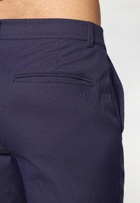 Close-up of a person wearing dark navy pants with a back welt pocket and a subtle embossed circular logo near the pocket.