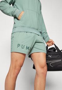 Bermudas deportivas verde claro con diseño del logo de PUMA, combinadas con una sudadera con capucha a juego. Se sostiene una bolsa de viaje negra en una mano.