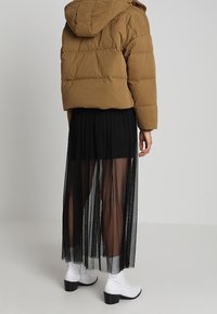 Personne portant une doudoune marron, une jupe noire transparente par-dessus un short, et des bottines blanches à talons, debout devant un fond uni.