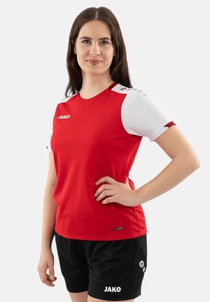 Frau trägt ein rotes Sportshirt mit weißen Ärmeln und schwarzen Shorts, steht mit einer Hand auf der Hüfte und lächelt leicht.