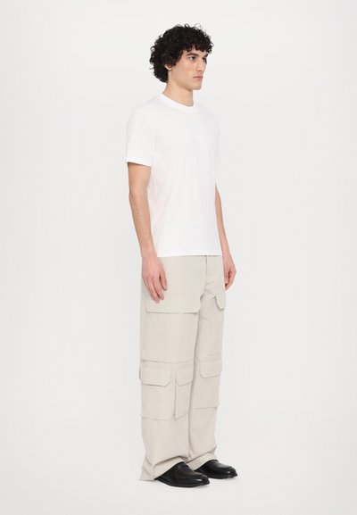 Tricou alb cu maneci scurte, purtat cu pantaloni scurți bej deschis, care au mai multe buzunare și o croială relaxată. Pantofi negri completează ținuta.