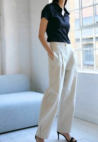 Chemise noire à col, à manches courtes, associée à un pantalon beige taille haute avec plis et poches latérales, complétée par des sandales à talons noires.