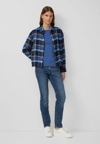 Junge Frau, die ein blaues Karohemd über einem blauen T-Shirt, enge blaue Jeans und weiße Sneakers trägt, steht vor einer schlichten hellgrauen Wand.
