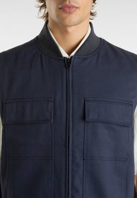 Gilet blu navy con colletto alto, chiusura frontale con zip e due tasche sul petto. Realizzato in tessuto resistente con finitura testurizzata.