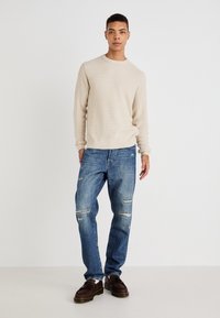 Beige gebreide trui met geribbeld uiteinde, gecombineerd met blauwe versleten jeans met kniebreuken. Bruine schoenen met een dikke zool maken de look compleet.
