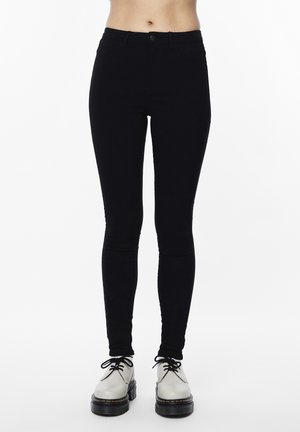 Czarne spodnie o dopasowanym kroju noszone przez stojącą osobę, zestawione z białymi platformowymi butami na sznurowadła na białym tle.