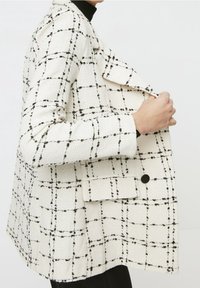 Blazer bianco strutturato con pattern a quadri neri, colletto a revers e chiusura con bottone. Il tessuto sembra intrecciato e ben definito.