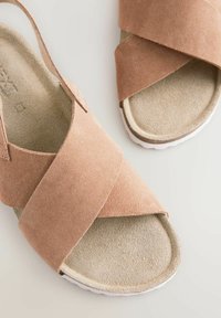 Sandales en daim tan avec des straps croisées et un coussin beige doux. Elles possèdent une semelle en caoutchouc blanche, conçue pour le confort et les tenues décontractées.