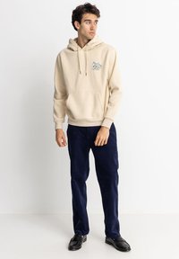 Sudadera beige con cordones y bolsillo delantero, con un diseño gráfico. Combinada con pantalones de pana azul marino y zapatos negros.