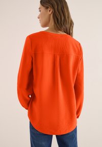 Femme aux longs cheveux bruns portant un chemisier à manches longues orange vif et un jean bleu, vue de dos sur un fond uni.