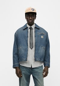 Νεαρός άνδρας με μπεζ καπέλο, υπερμεγέθη μπλε τζιν μπουφάν, λευκό πουκάμισο, μαύρη γραβάτα και μπλε τζιν παντελόνι, στέκεται μπροστά σε απλό λευκό φόντο.