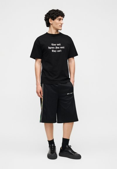 Νέος άνδρας φοράει μαύρο T-shirt με λευκό κείμενο «They will Ignore You until They can’t» και μαύρα σορτς με ρίγες στο πλάι.