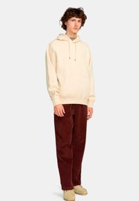 Sudadera de color beige claro con un bolsillo frontal y cordón, combinada con pantalones de pana burdeos oscuro. Diseño minimalista y corte relajado.