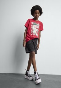 Červené tričko s grafickým potiskem, černé džíny šortky a černé vysoké tenisky Converse s bílými tkaničkami, stojící na šedé podlaze.