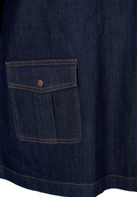 Tummah denim-kangas sisältää suuren läppätaskun, jossa on kupari-napsulukko, ja jota korostaa kontrastivärinen oranssi tikkaus.