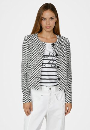 Frau trägt eine schwarz-weiß gemusterte Jacke mit Knöpfen über einem gestreiften Hemd mit Text, kombiniert mit hoch taillierten weißen Hosen.