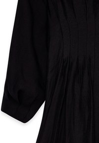 Juoda suknelė su ilgomis, išpūstomis rankovėmis; turi vertikalių raukšlių ir lygų, tekstūruotą audinį. Paprasta, elegantiška siluetas.