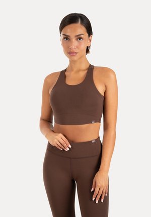 Frau trägt einen braunen Sport-BH und passende Leggings mit hoher Taille, steht mit einer Hand in der Taille vor einem schlichten Hintergrund.