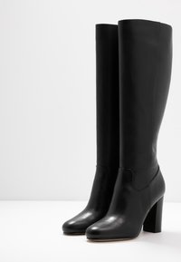 Dvojice černých kožených vysokých bot po kolena se zaoblenými špičkami a blokovými podpatky na bílé ploše na jednobarevném pozadí.